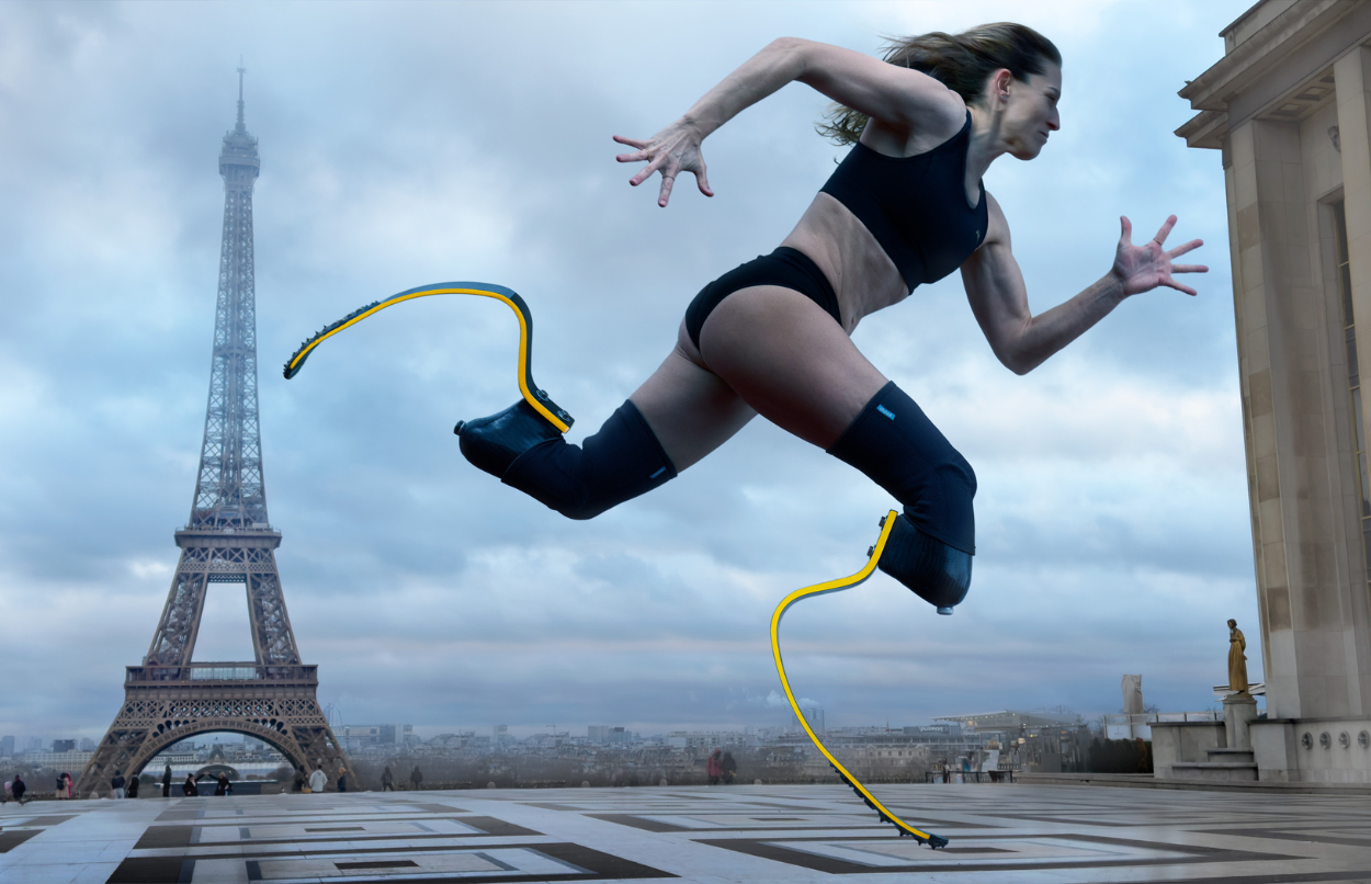 Para Athlete Sara Andrés Barrio with running blades is mid-stride in front of the Eiffel Tower. She wears a sports bra and shorts. The background features a cloudy sky and Parisian architecture.